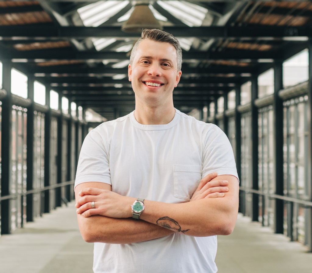 A photo of Alex Marcus standing in a hallway while wearing a white shirt and smiling.